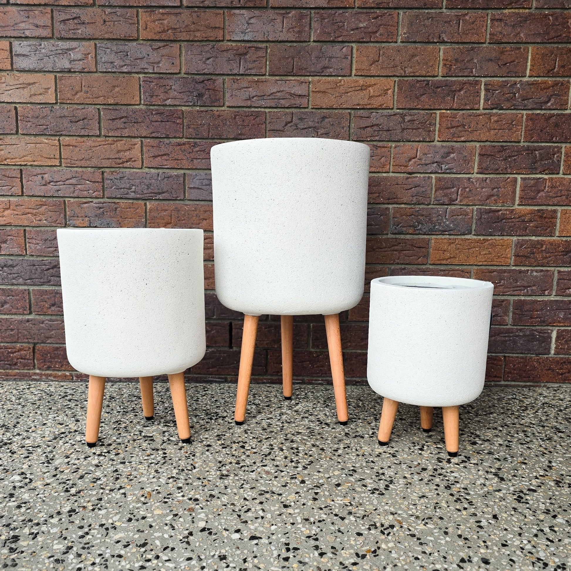 Vellum Tripod Pots Indoor White Pots on wooden legs, available in small, medium and large as showing three different sizes on the pictures, brick wall behind on the concrete floor. Brisbane Indoor Pots Delivered to your door. 